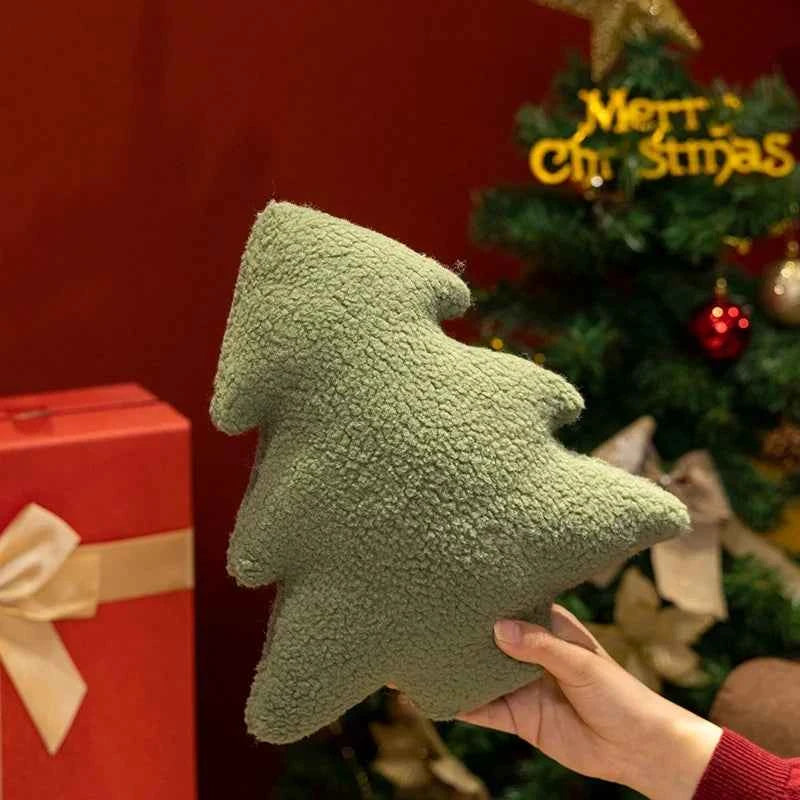 Green tree-shaped plush toy held in front of a Christmas tree and gift box.